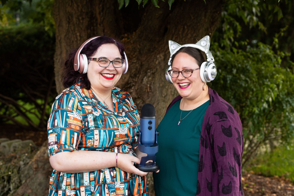 Kait and Renata, posed outside wearing headphones and holding a microphone