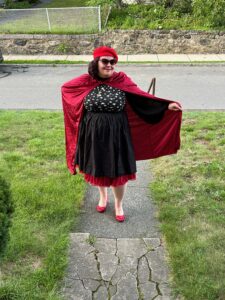 Kait posing dramatically with her poetry outfit: a black dress, red beret, red cape, and sunglasses